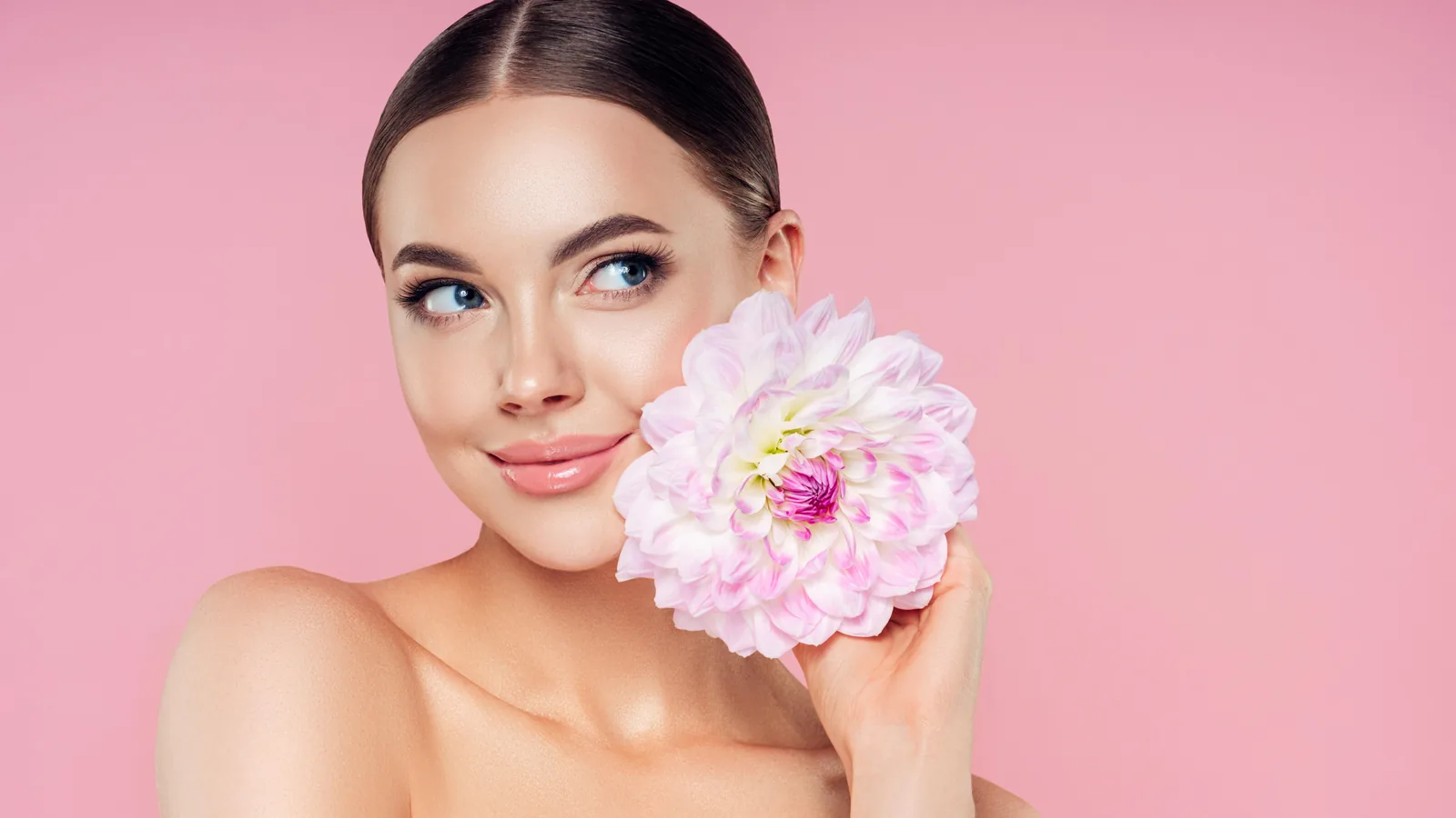 Woman holding a pale pink peony beside her face on a pink background